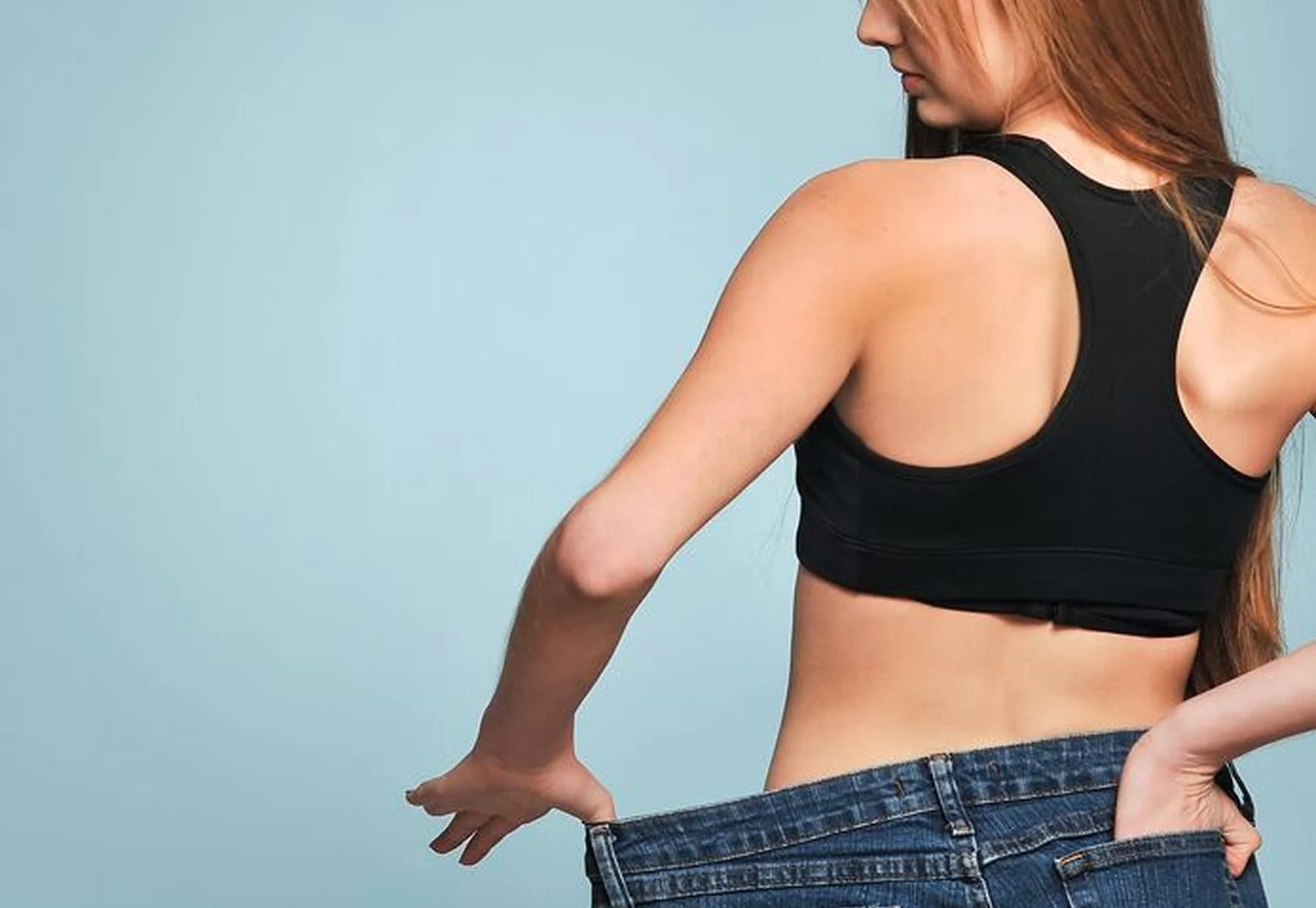 A young woman holds out the waistband of a loose pair of jeans, indicating successful weight loss.